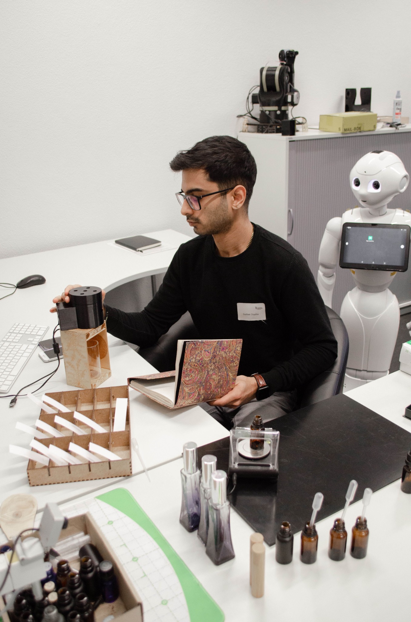 Sarthak Chadha working with an olfactory prototype at a lab table, with a Pepper robot beside him